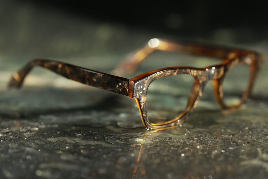 Handgefertigte Designerbrille mit einzigartiger Baumwollacetatstruktur – ein stilvolles Unikat für Damen und Herren aus der Brillenmanufaktur Hafenoptiker Detailansicht von vorne-rechts einer rechteckigen Brillenfassung aus mehrfarbig gemasertem Baumwollacetat, die auf einem Tisch mit reflektierender Oberfläche liegt