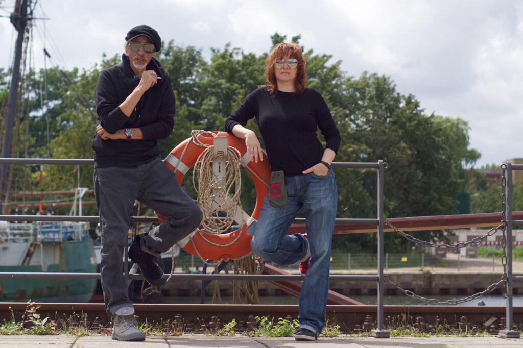 Die kreativen Köpfe hinter der Brillenmanufaktur Hafenoptiker – stilvoll und authentisch am Hafen, wo Handwerk auf Charakter trifft Bruecher und Taege, die Inhaber der Brillenmanufaktur Hafenoptiker, posieren an einem Geländer mit Rettungsring vor einem Hafen im Hintergrund.